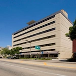 A beige multi-story office building with large windows stands beside a quiet, sunlit street in Lincoln, Nebraska, lined with trees and signs under a clear blue sky—an inviting setting near Crossroads House for independent living.