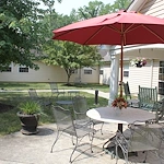 Outdoor sitting area with table, umbrella, and chairs on a cement pad surrounded by green grass, trees, and a sidewalk at Charter Senior Living of Jackson in Jackson, TN, providing a peaceful space for assisted living and memory care residents.