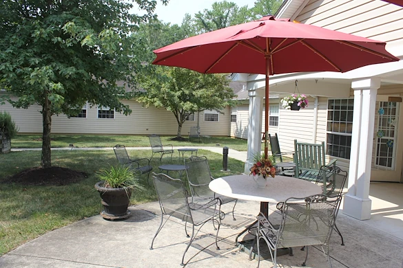 Outdoor sitting area with table, umbrella, and chairs on a cement pad surrounded by green grass, trees, and a sidewalk at Charter Senior Living of Jackson in Jackson, TN, providing a peaceful space for assisted living and memory care residents.