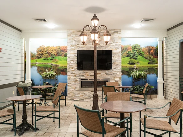 Comfortable common area at Charter Senior Living of Hazel Crest in Hazel Crest, IL, featuring a fireplace with natural landscape wallpaper, TV, chairs, tables, and tile flooring, designed for relaxation and socialization in assisted living and memory care.