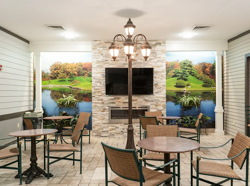 Comfortable common area at Charter Senior Living of Hazel Crest in Hazel Crest, IL, featuring a fireplace with natural landscape wallpaper, TV, chairs, tables, and tile flooring, designed for relaxation and socialization in assisted living and memory care.