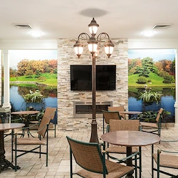 Comfortable common area at Charter Senior Living of Hazel Crest in Hazel Crest, IL, featuring a fireplace with natural landscape wallpaper, TV, chairs, tables, and tile flooring, designed for relaxation and socialization in assisted living and memory care.