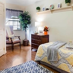 Inviting assisted living and memory care bedroom at Charter Senior Living of Jackson in Jackson, TN, featuring a comfortable bed, dresser, nightstand, cozy corner chair, and a large window letting in natural light for a warm, safe environment.