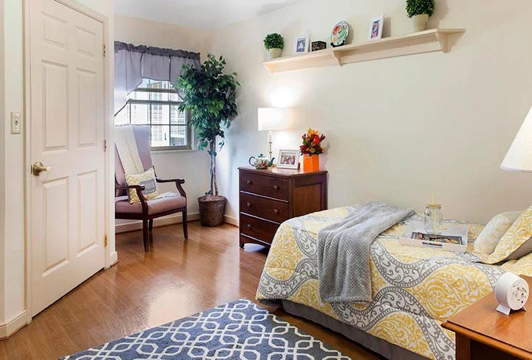 Inviting assisted living and memory care bedroom at Charter Senior Living of Jackson in Jackson, TN, featuring a comfortable bed, dresser, nightstand, cozy corner chair, and a large window letting in natural light for a warm, safe environment.