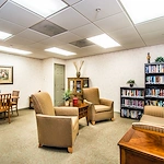 A bright, cozy room in Godfrey, Illinois features a small wooden dining table with four chairs, two armchairs, a striped loveseat, bookshelves filled with books—an inviting space ideal for assisted living or memory care residents.