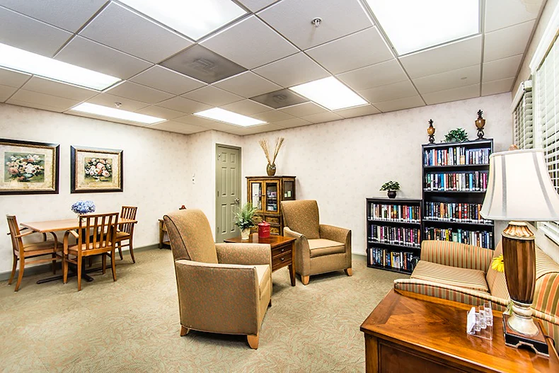 A bright, cozy room in Godfrey, Illinois features a small wooden dining table with four chairs, two armchairs, a striped loveseat, bookshelves filled with books—an inviting space ideal for assisted living or memory care residents.