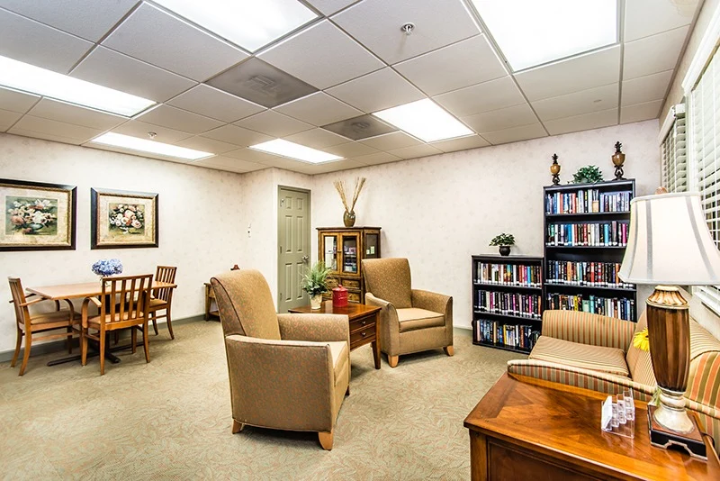 A bright, cozy room in Godfrey, Illinois features a small wooden dining table with four chairs, two armchairs, a striped loveseat, bookshelves filled with books—an inviting space ideal for assisted living or memory care residents.