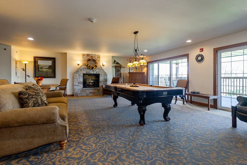 A cozy game room in Jackson, Wisconsin, perfect for assisted living and memory care, features a pool table at the center, beige sofas, a stone fireplace with a painting above, natural light from large windows, and ornate lighting over patterned blue carpet.