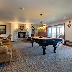 A cozy game room in Jackson, Wisconsin, perfect for assisted living and memory care, features a pool table at the center, beige sofas, a stone fireplace with a painting above, natural light from large windows, and ornate lighting over patterned blue carpet.