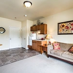 This cozy assisted living apartment in Godfrey, Illinois, features a beige sofa with floral pillows, a wooden dining table, a small kitchen with microwave and cabinets, and floral artwork above the couch. Flooring transitions from tile to carpet for comfort.