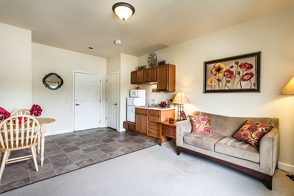 This cozy assisted living apartment in Godfrey, Illinois, features a beige sofa with floral pillows, a wooden dining table, a small kitchen with microwave and cabinets, and floral artwork above the couch. Flooring transitions from tile to carpet for comfort.