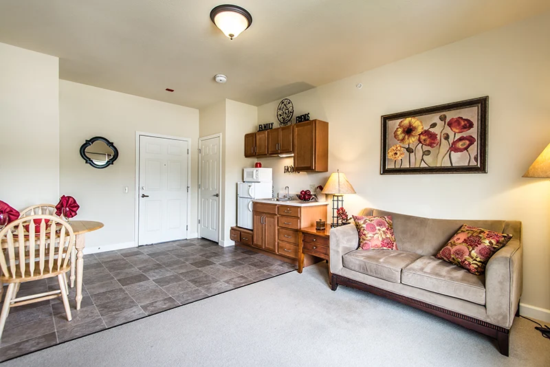 This cozy assisted living apartment in Godfrey, Illinois, features a beige sofa with floral pillows, a wooden dining table, a small kitchen with microwave and cabinets, and floral artwork above the couch. Flooring transitions from tile to carpet for comfort.