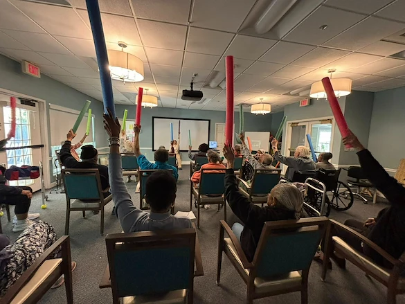 Seniors in assisted living and memory care at Charter Senior Living of Hazel Crest in Hazel Crest, IL, participating in an active group exercise with pool noodles, promoting physical activity and social engagement.