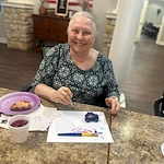 An older woman with short gray hair smiles at a table while painting a small wooden car in an assisted living setting. She wears a patterned top, with art supplies, pizza on a purple plate, and patriotic decor visible in the Godfrey, Illinois background.
