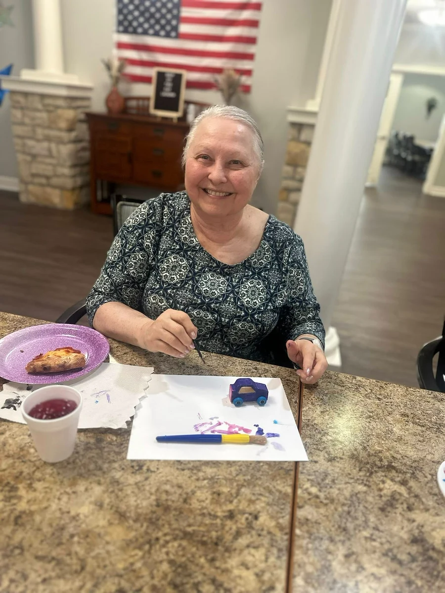 An older woman with short gray hair smiles at a table while painting a small wooden car in an assisted living setting. She wears a patterned top, with art supplies, pizza on a purple plate, and patriotic decor visible in the Godfrey, Illinois background.