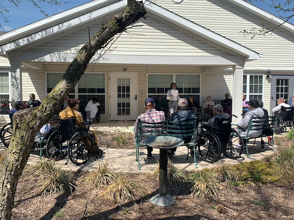 Residents at Charter Senior Living of Hazel Crest in Hazel Crest, IL, gathered outdoors as someone reads aloud, enjoying a peaceful moment and shared engagement in assisted living and memory care.