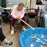 Resident at Charter Senior Living of Jackson in Jackson, TN, enjoying a fun fishing activity using a wooden pole to catch paper fish from a blue plastic pool, promoting engagement and recreation in assisted living and memory care.