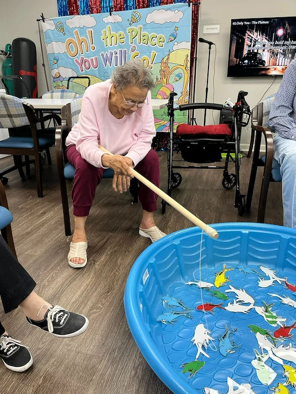 Resident at Charter Senior Living of Jackson in Jackson, TN, enjoying a fun fishing activity using a wooden pole to catch paper fish from a blue plastic pool, promoting engagement and recreation in assisted living and memory care.
