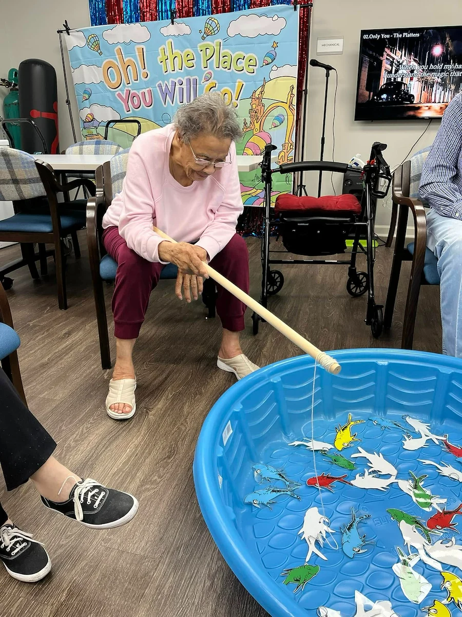 Resident at Charter Senior Living of Jackson in Jackson, TN, enjoying a fun fishing activity using a wooden pole to catch paper fish from a blue plastic pool, promoting engagement and recreation in assisted living and memory care.