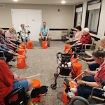 In an assisted living center in Godfrey, Illinois, a group of elderly people sit in a circle, drumming on orange Home Depot buckets. Some use walkers or wheelchairs, while a man leads the engaging and communal session.