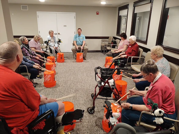 In an assisted living center in Godfrey, Illinois, a group of elderly people sit in a circle, drumming on orange Home Depot buckets. Some use walkers or wheelchairs, while a man leads the engaging and communal session.