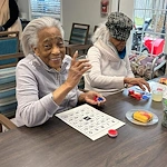 Resident at Charter Senior Living of Hazel Crest in Hazel Crest, IL, smiling while playing Bingo and enjoying a sweet treat at a table in the main common area, showcasing joy and engagement in assisted living and memory care.