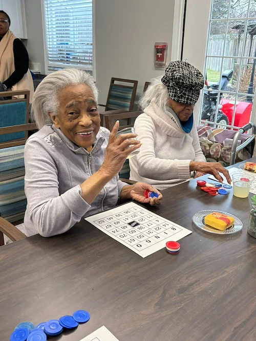 Resident at Charter Senior Living of Hazel Crest in Hazel Crest, IL, smiling while playing Bingo and enjoying a sweet treat at a table in the main common area, showcasing joy and engagement in assisted living and memory care.