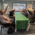 Residents at Charter Senior Living of Hazel Crest in Hazel Crest, IL, celebrating a football-themed Super Bowl gathering with hands in the air, enjoying the game on a large screen TV in the assisted living and memory care common area.