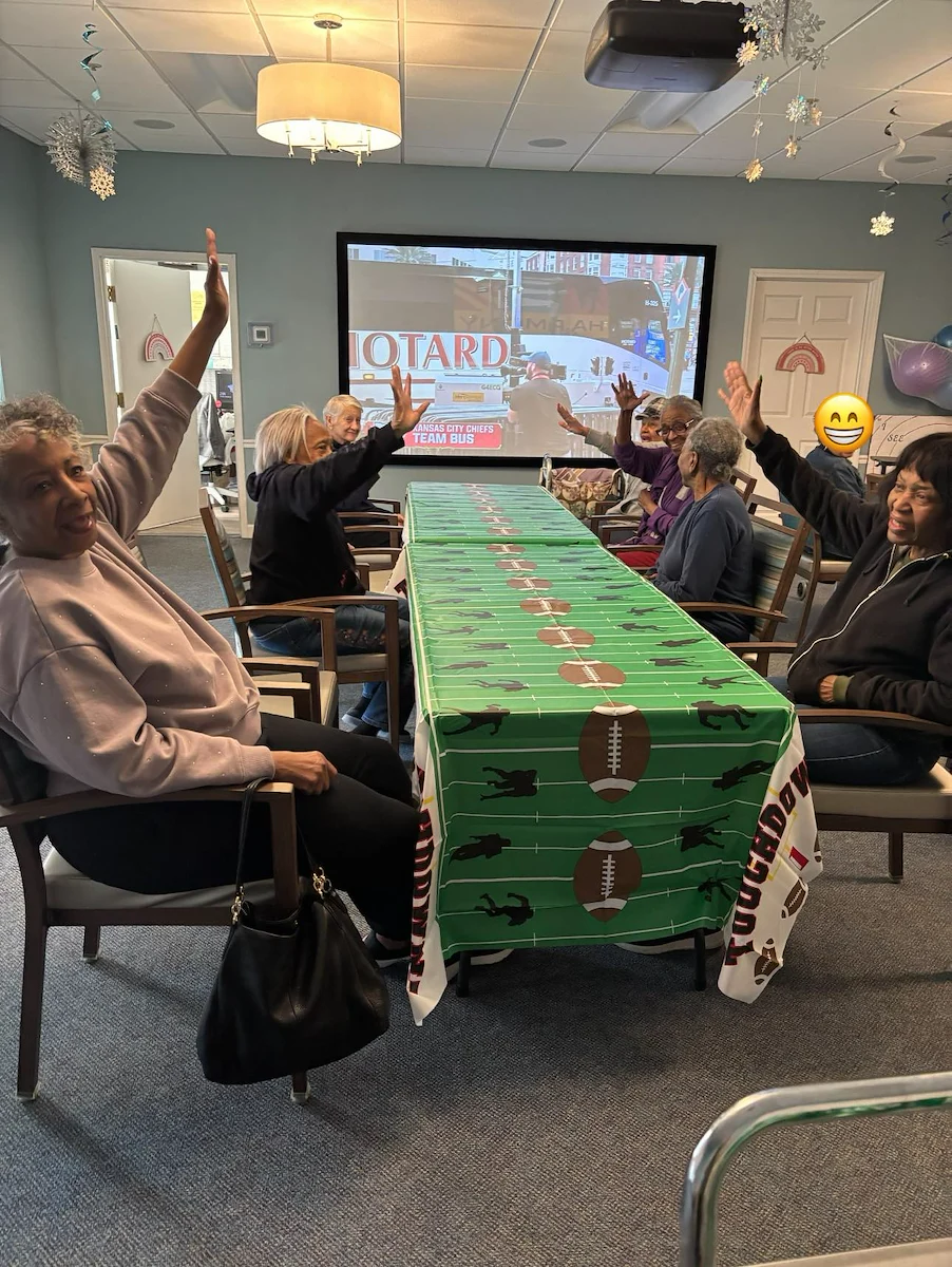 Residents at Charter Senior Living of Hazel Crest in Hazel Crest, IL, celebrating a football-themed Super Bowl gathering with hands in the air, enjoying the game on a large screen TV in the assisted living and memory care common area.