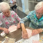 Two residents at Charter Senior Living of Hopkinsville in Hopkinsville, KY, engaged in a creative craft activity using decorative paper, lace, and pins, fostering artistic expression and social engagement in assisted living and memory care.
