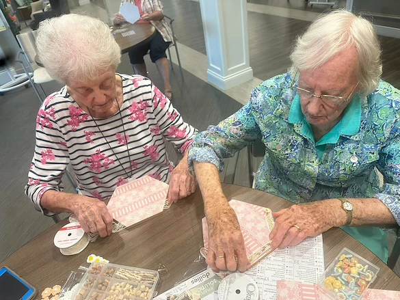 Two residents at Charter Senior Living of Hopkinsville in Hopkinsville, KY, engaged in a creative craft activity using decorative paper, lace, and pins, fostering artistic expression and social engagement in assisted living and memory care.