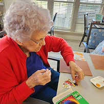 An elderly woman with curly gray hair and glasses sits at a table in Charter Senior Living Gallatin, assembling a jigsaw puzzle. She is wearing a red cardigan and blue shirt, enjoying assisted living in a bright room with large windows in the background.