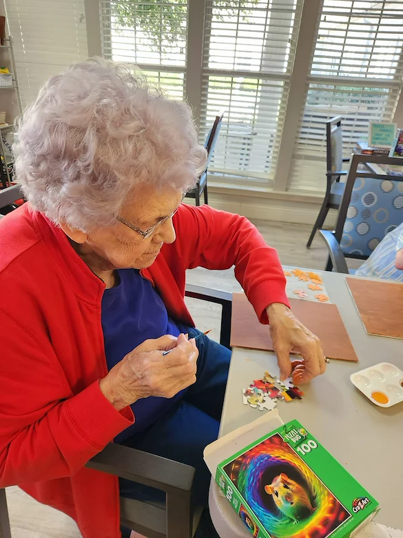An elderly woman with curly gray hair and glasses sits at a table in Charter Senior Living Gallatin, assembling a jigsaw puzzle. She is wearing a red cardigan and blue shirt, enjoying assisted living in a bright room with large windows in the background.