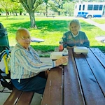 Two elderly women sit at a wooden picnic table outdoors in Godfrey, Illinois, enjoying a meal from takeout containers under a shaded pavilion—an inviting moment that reflects the warm community feel of local assisted living.