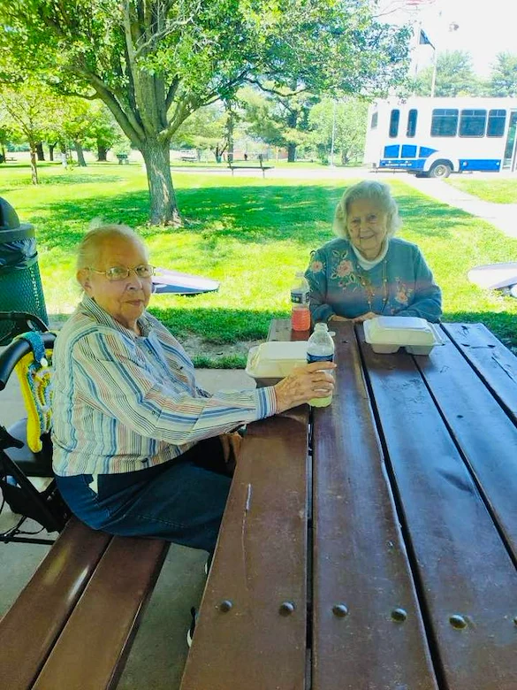 Two elderly women sit at a wooden picnic table outdoors in Godfrey, Illinois, enjoying a meal from takeout containers under a shaded pavilion—an inviting moment that reflects the warm community feel of local assisted living.