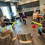 Seniors at Charter Senior Living of Hopkinsville in Hopkinsville, KY, sitting in a circle engaging in an active group activity with pool noodles and a balloon, promoting fun, fitness, and community in assisted living and memory care.
