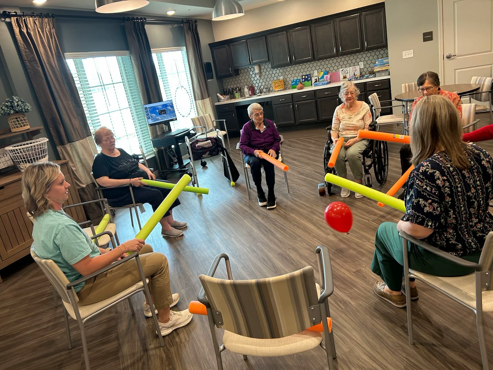 Seniors at Charter Senior Living of Hopkinsville in Hopkinsville, KY, sitting in a circle engaging in an active group activity with pool noodles and a balloon, promoting fun, fitness, and community in assisted living and memory care.