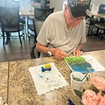 An elderly man in a gray shirt and black cap paints at a table in a Godfrey, Illinois assisted living community. A small toy car and art supplies are before him, with flowers and soft natural light filling the room from large windows.