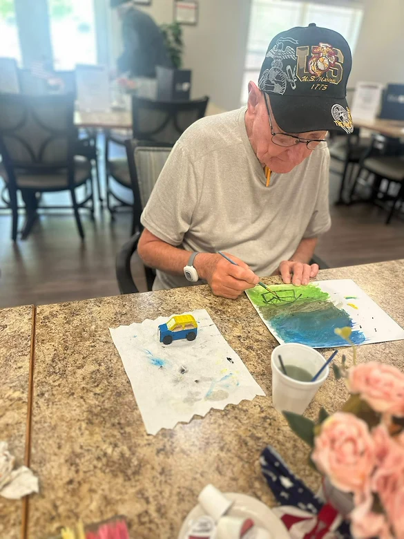 An elderly man in a gray shirt and black cap paints at a table in a Godfrey, Illinois assisted living community. A small toy car and art supplies are before him, with flowers and soft natural light filling the room from large windows.