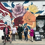 Five older women stand smiling in front of a vibrant mural with colorful flowers on a white wall in Godfrey, Illinois. One woman uses a red walker. A black sign reads NOW OPEN THE CURIOUS WILSON—an inviting spot for assisted living and memory care.