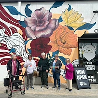 Five older women stand smiling in front of a vibrant mural with colorful flowers on a white wall in Godfrey, Illinois. One woman uses a red walker. A black sign reads NOW OPEN THE CURIOUS WILSON—an inviting spot for assisted living and memory care.