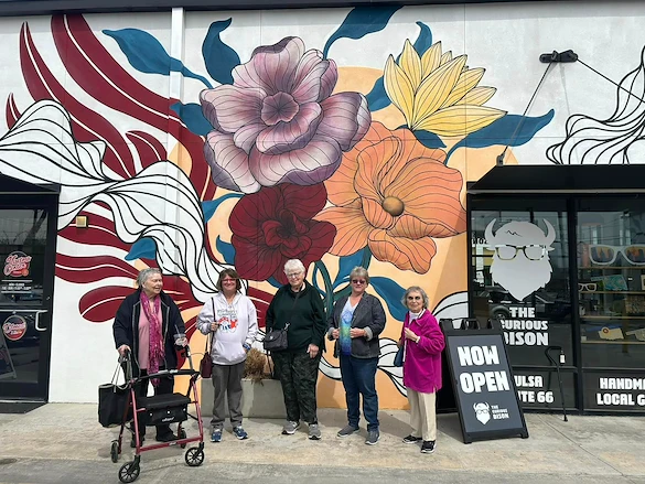Five older women stand smiling in front of a vibrant mural with colorful flowers on a white wall in Godfrey, Illinois. One woman uses a red walker. A black sign reads NOW OPEN THE CURIOUS WILSON—an inviting spot for assisted living and memory care.