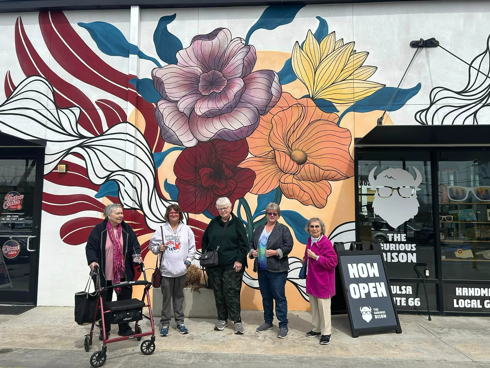 Five older women stand smiling in front of a vibrant mural with colorful flowers on a white wall in Godfrey, Illinois. One woman uses a red walker. A black sign reads NOW OPEN THE CURIOUS WILSON—an inviting spot for assisted living and memory care.