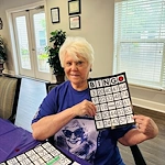 An elderly woman with short white hair and a purple shirt smiles while holding up a winning bingo card at an assisted living community in Godfrey, Illinois. Bingo chips, papers, and a pen are on the table; large windows and plants brighten the cozy room.