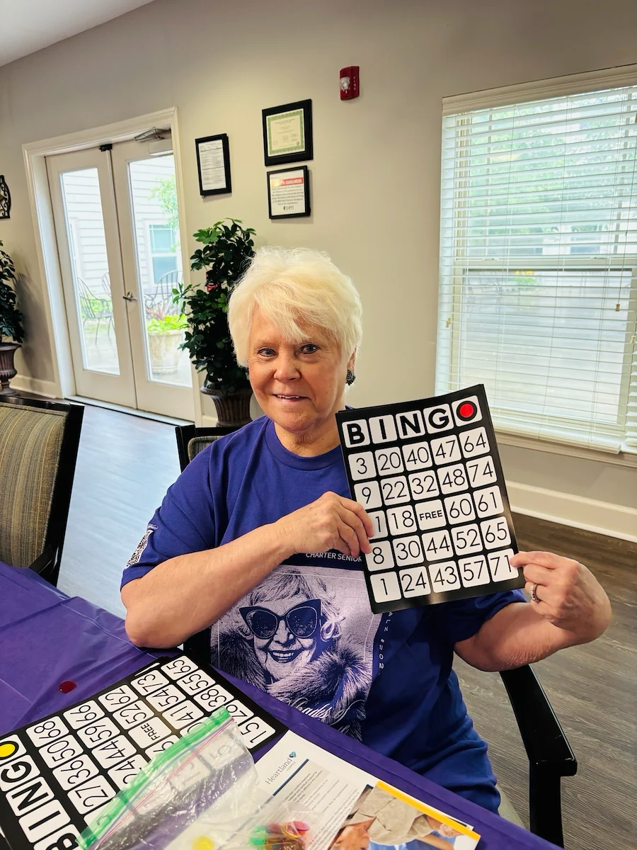 An elderly woman with short white hair and a purple shirt smiles while holding up a winning bingo card at an assisted living community in Godfrey, Illinois. Bingo chips, papers, and a pen are on the table; large windows and plants brighten the cozy room.