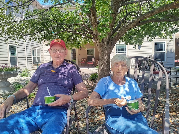 An elderly man and woman sit outdoors on metal chairs under a leafy tree, enjoying green bowls of ice cream at an assisted living community in Godfrey, Illinois. They wear casual clothes and glasses, appearing relaxed and content in the sunny garden.