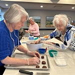 Two elderly women wearing gloves shape and place chocolate cookies on a baking tray in a bright kitchen at an assisted living community in Godfrey, Illinois. Other seniors work in the background, with bowls and utensils scattered across the counter.