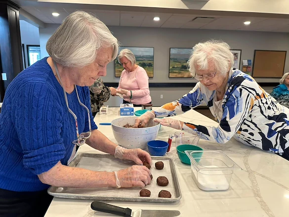 Two elderly women wearing gloves shape and place chocolate cookies on a baking tray in a bright kitchen at an assisted living community in Godfrey, Illinois. Other seniors work in the background, with bowls and utensils scattered across the counter.