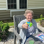 Smiling resident at Charter Senior Living of Jackson in Jackson, TN, enjoying outdoor fun with a bubble machine while sitting at a table in the sunshine, promoting happiness and wellness in assisted living and memory care.