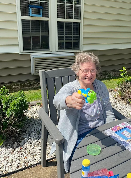 Smiling resident at Charter Senior Living of Jackson in Jackson, TN, enjoying outdoor fun with a bubble machine while sitting at a table in the sunshine, promoting happiness and wellness in assisted living and memory care.